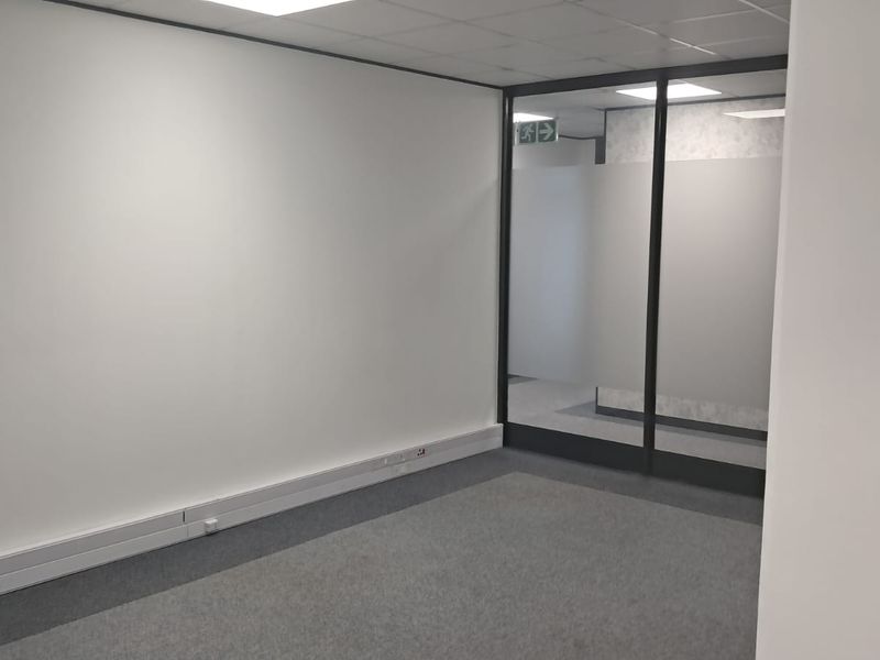 A clean, empty office space featuring neutral walls, carpeted flooring, a suspended ceiling, and a modern glass partition with frosted privacy film.