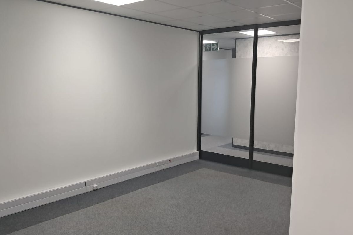 A clean, empty office space featuring neutral walls, carpeted flooring, a suspended ceiling, and a modern glass partition with frosted privacy film.
