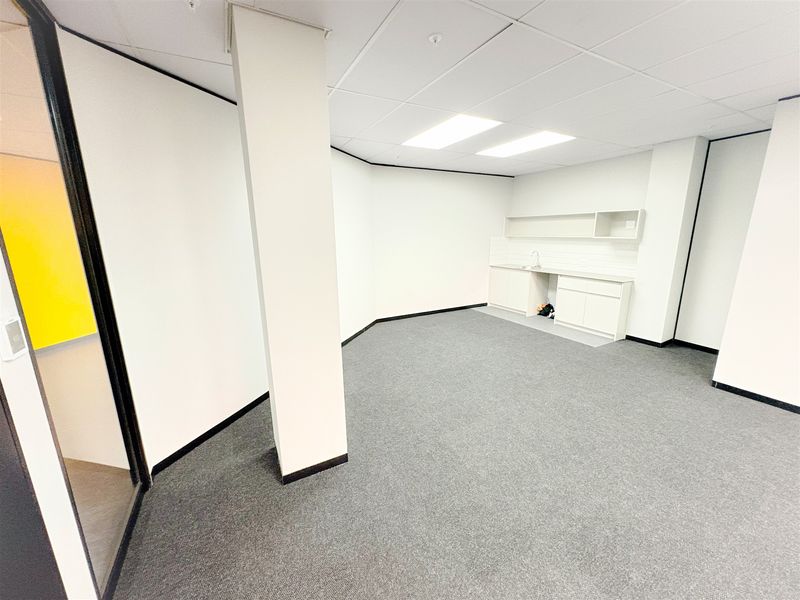 A clean, vacant office space featuring a built-in kitchenette, neutral carpet flooring, and a suspended ceiling with integrated lighting.
