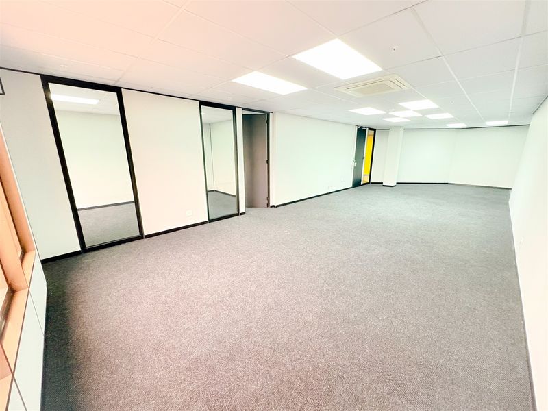 A spacious, empty office area featuring neutral carpet flooring, a suspended ceiling with recessed lighting, and integrated mirror panels.