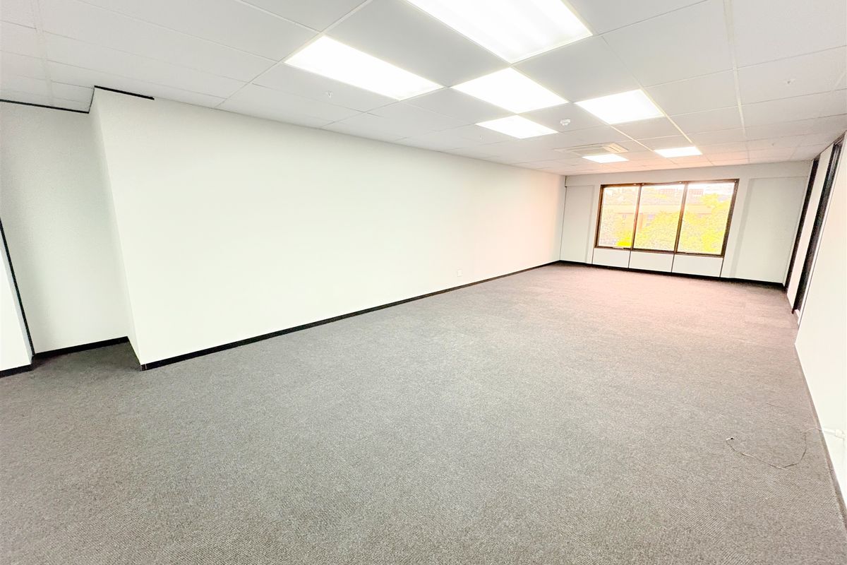 A spacious, empty office suite featuring neutral carpeted flooring, a clean suspended ceiling with recessed lighting, and large windows providing natural light.