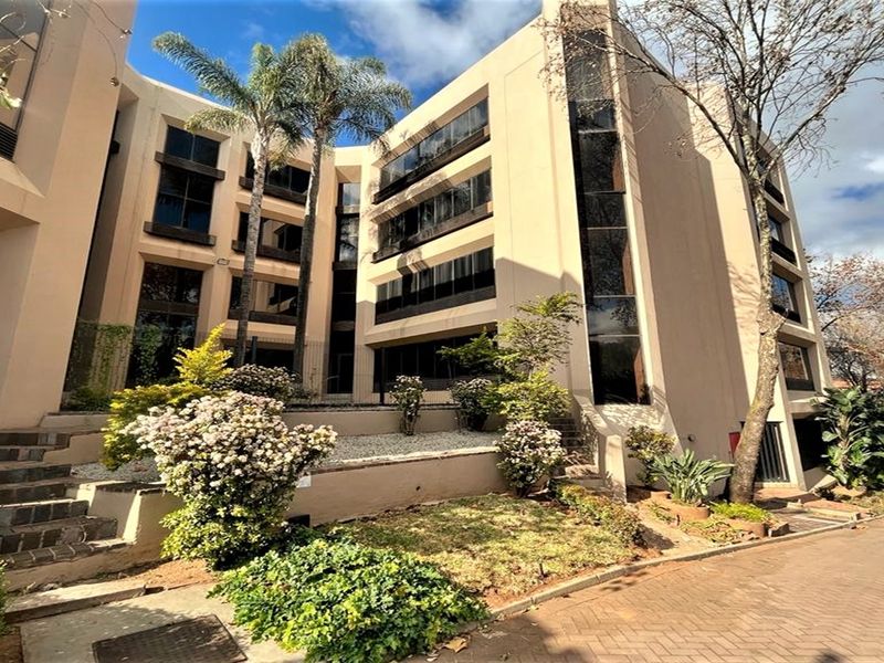 A multi-story office building facade featuring tinted glass windows, paved driveway access, and established landscaped garden beds.