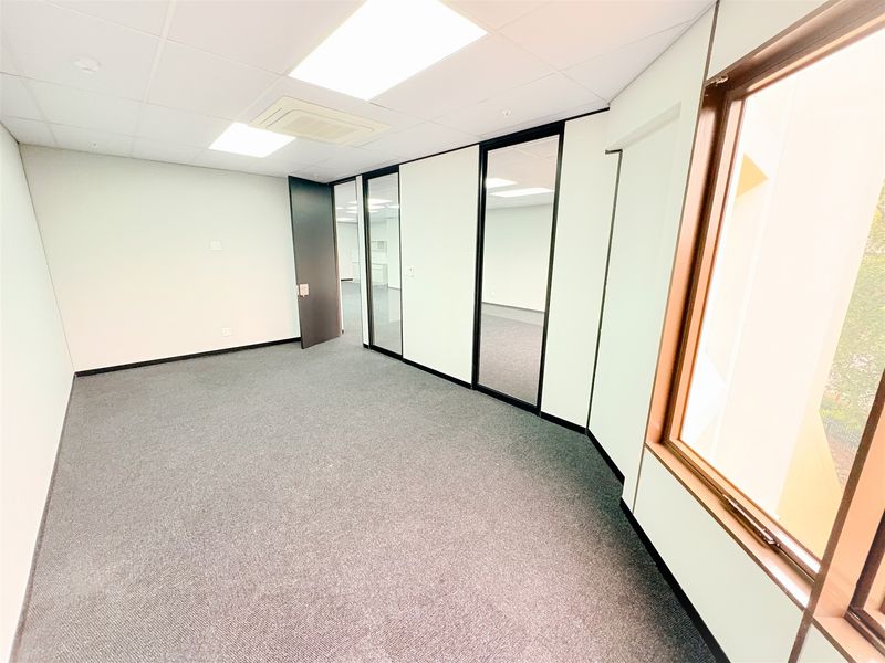 A bright, empty office space featuring carpeted flooring, glass partitions, and a suspended ceiling with integrated lighting and air conditioning.