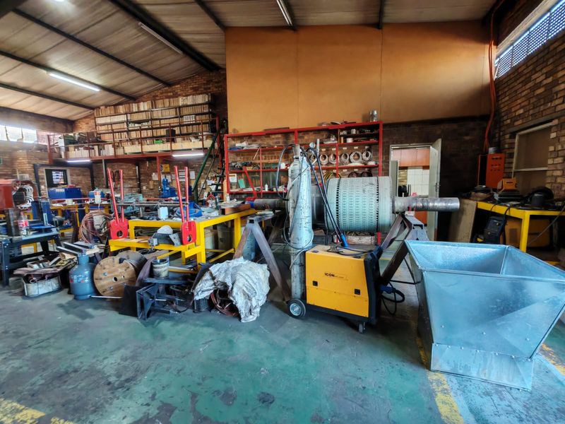 Industrial workshop space featuring exposed brick walls, high-clearance steel roof trusses, concrete flooring, and integrated mezzanine storage shelving.