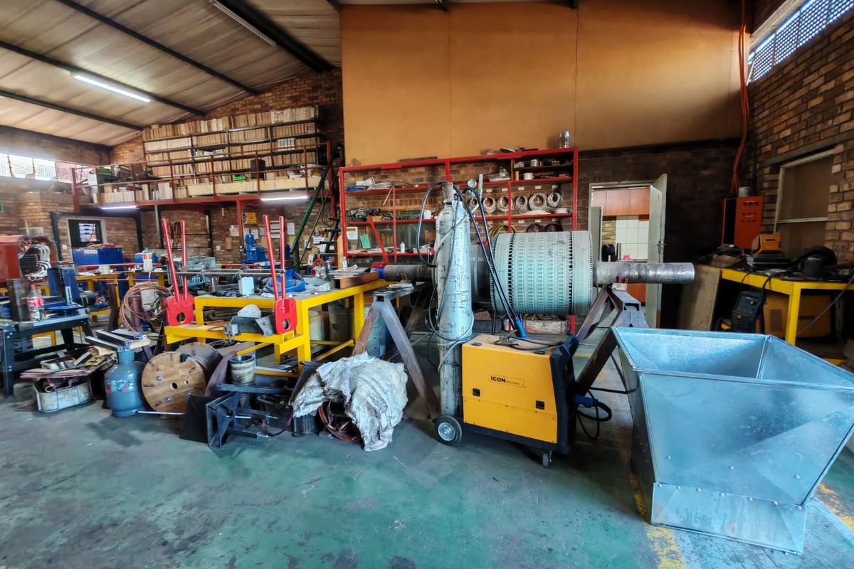Industrial workshop space featuring exposed brick walls, high-clearance steel roof trusses, concrete flooring, and integrated mezzanine storage shelving.