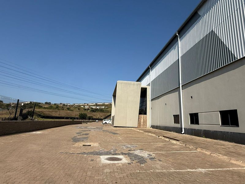 Exterior view of an industrial property featuring a large paved parking area, corrugated metal cladding, and a prominent concrete architectural entrance feature.