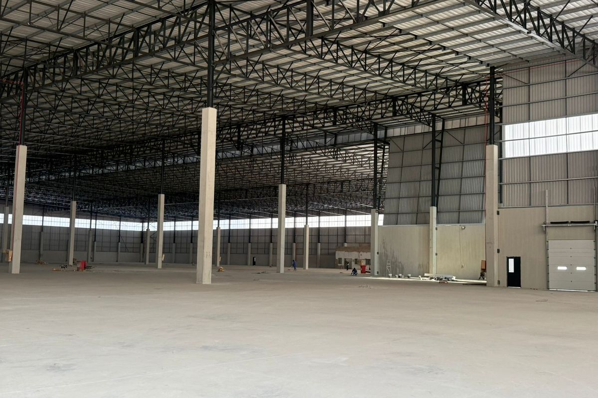 A large, open-plan industrial warehouse space featuring high ceilings with exposed steel trusses, polished concrete flooring, and a large roller shutter door.