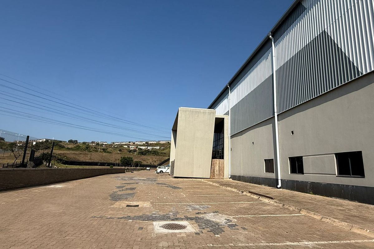 Exterior view of an industrial property featuring a large paved parking area, corrugated metal cladding, and a prominent concrete architectural entrance feature.