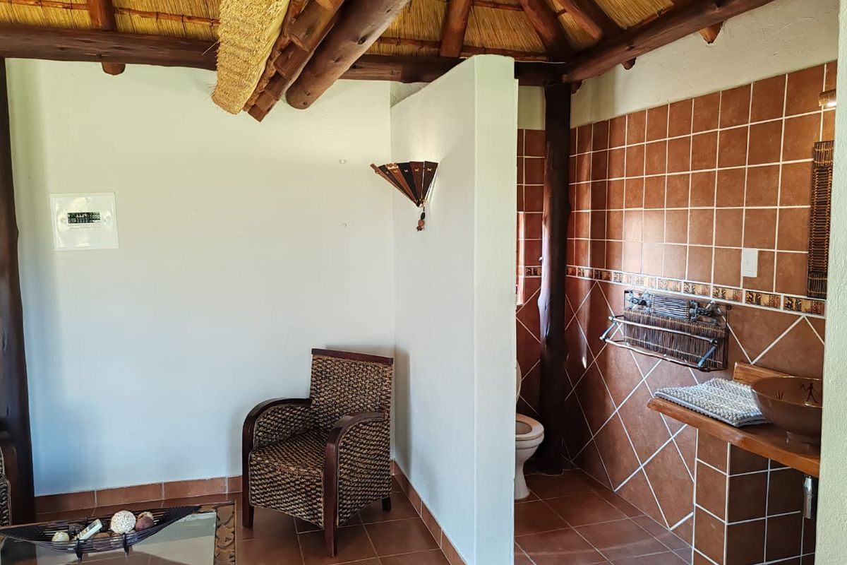 A rustic bathroom featuring a traditional thatch roof with exposed timber beams, terracotta-style wall and floor tiling, and a built-in vanity area.