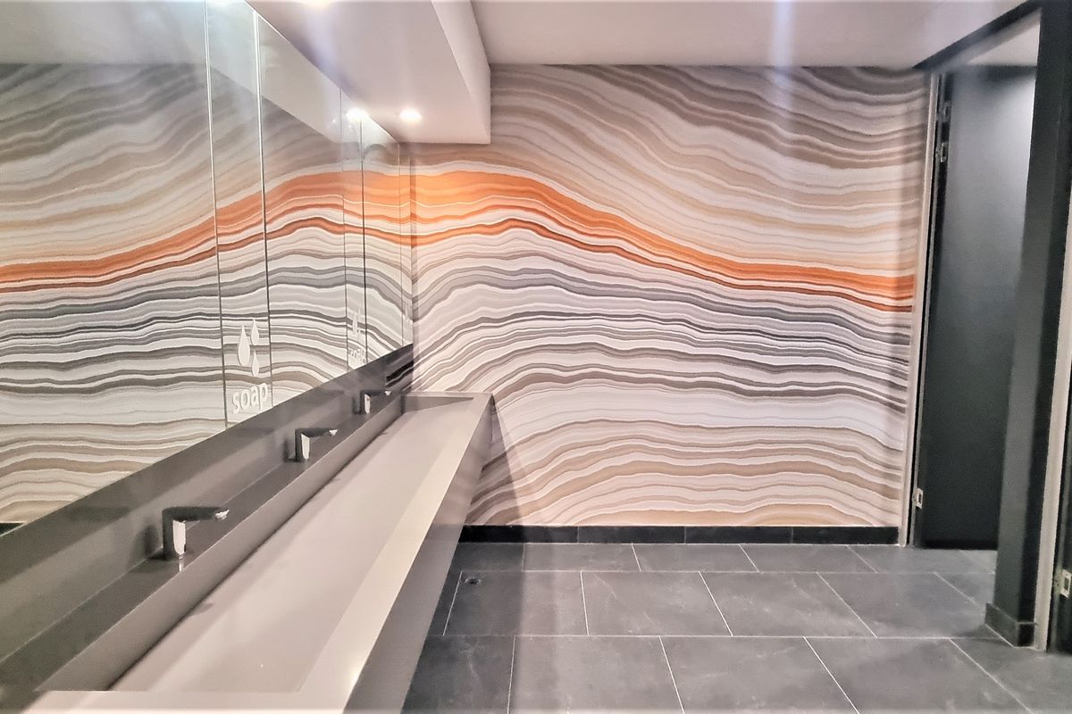 Modern office restroom featuring a long integrated trough sink, large wall-to-wall mirror, and decorative patterned feature wall with dark floor tiling.