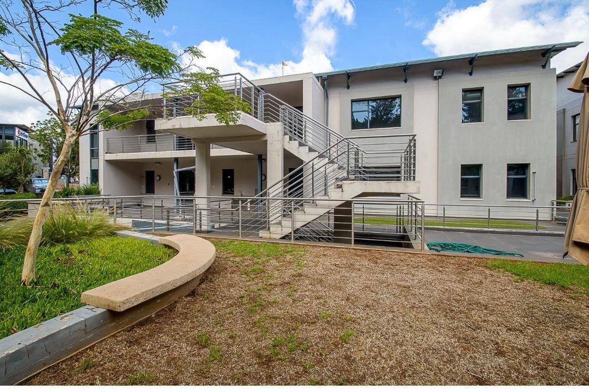 Rear view of the office building featuring an external concrete staircase with stainless steel railings, paved courtyard area, and landscaped garden beds.