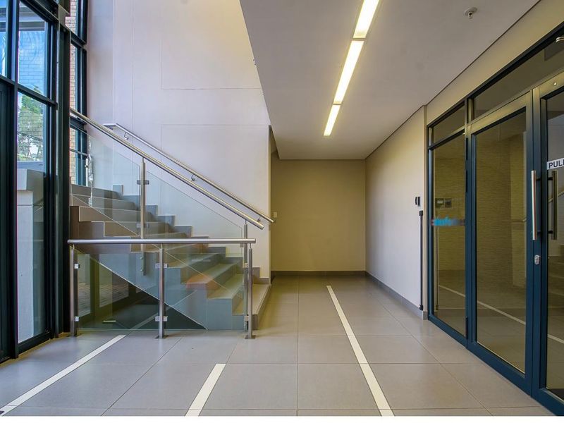 Modern office building entrance lobby featuring tiled flooring, glass balustrade staircase, and contemporary linear lighting with large glass entry doors.