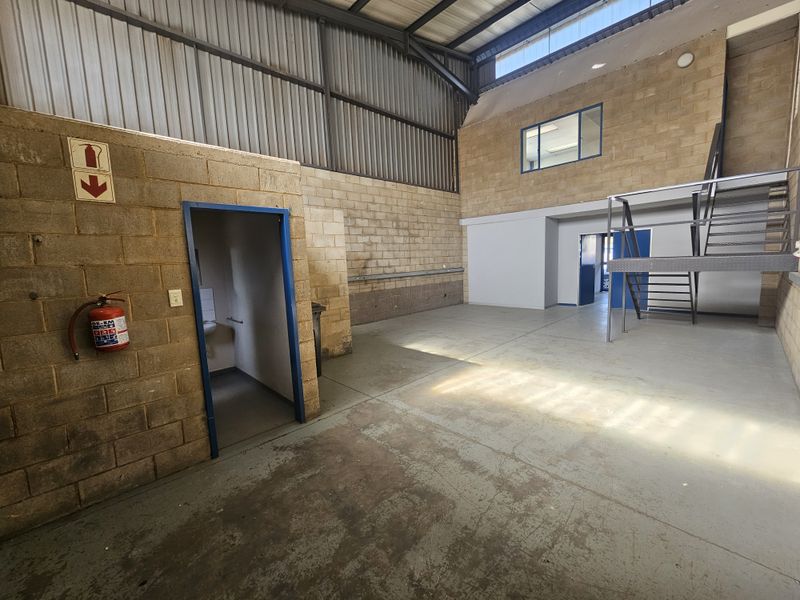 Spacious industrial utility area featuring exposed brick walls, concrete flooring, a metal staircase leading to a mezzanine level, and high-clearance steel roof trusses.