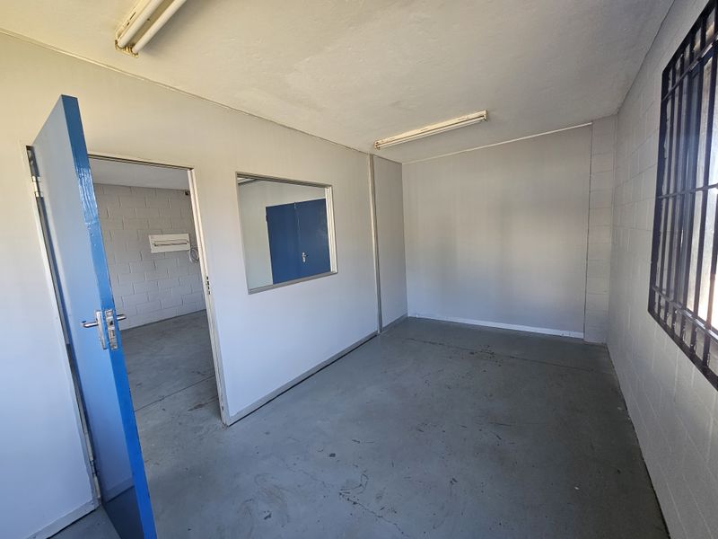 An empty industrial office space featuring painted blockwork walls, concrete flooring, security bars on windows, and fluorescent lighting.