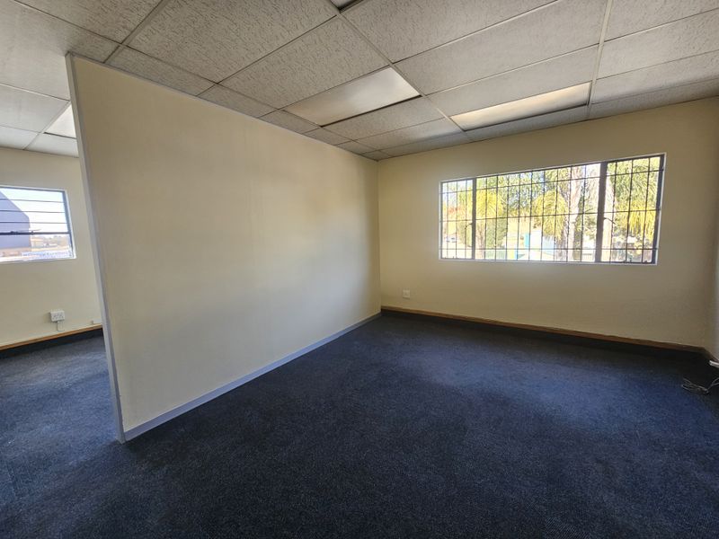 An empty office space featuring a suspended ceiling, recessed lighting, security bars on windows, and carpeted flooring.
