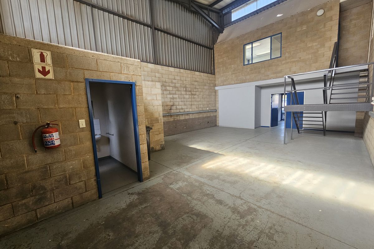 Spacious industrial utility area featuring exposed brick walls, concrete flooring, a metal staircase leading to a mezzanine level, and high-clearance steel roof trusses.