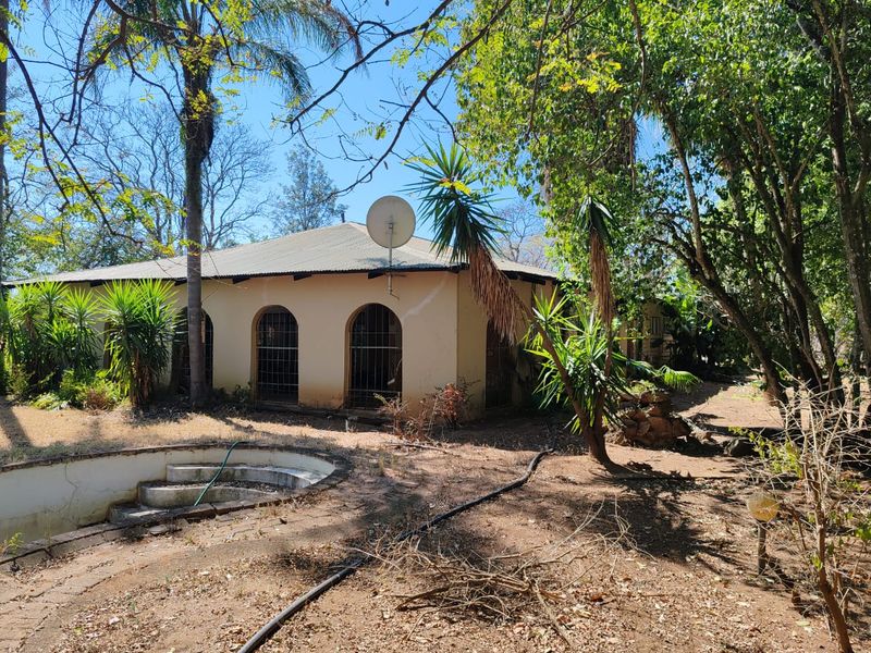 Rear view of a farm property featuring a swimming pool, arched windows with security bars, and a corrugated metal roof set within a wooded garden.