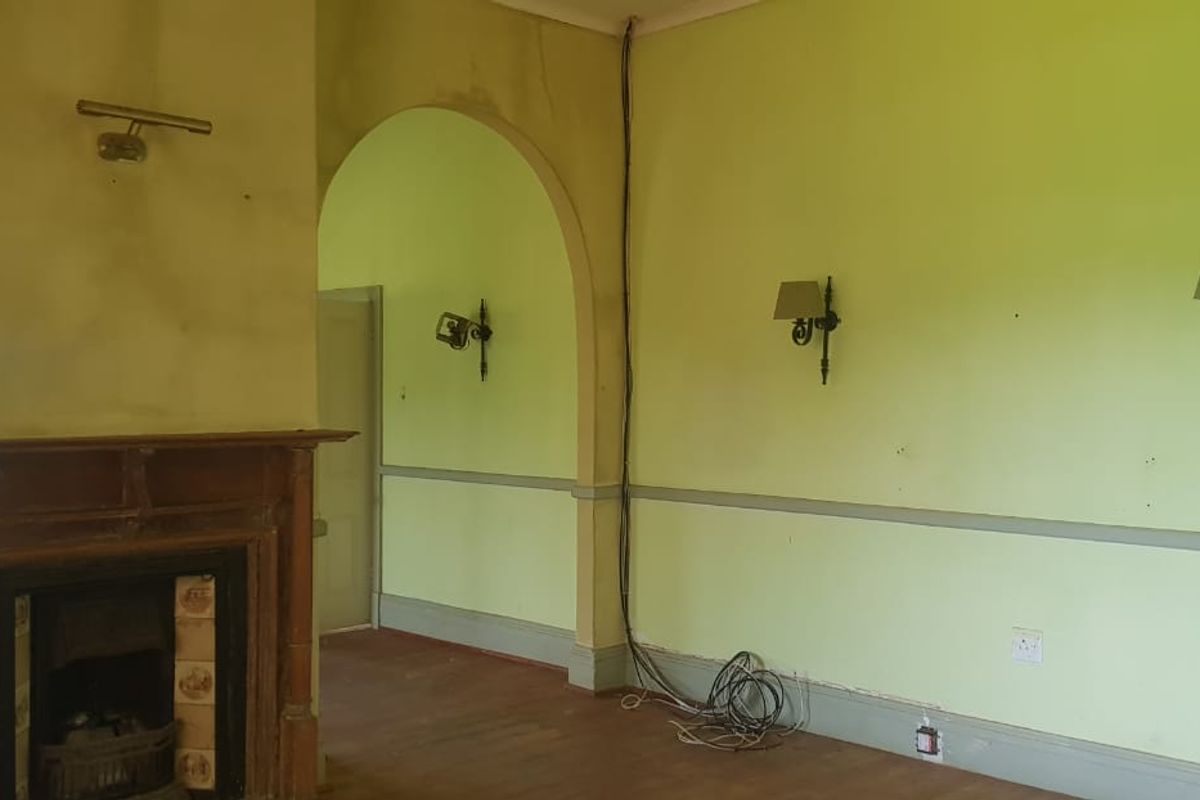 An empty living room featuring a traditional fireplace with a wooden mantelpiece, arched doorway, and wall sconces, requiring cosmetic renovation and electrical repairs.