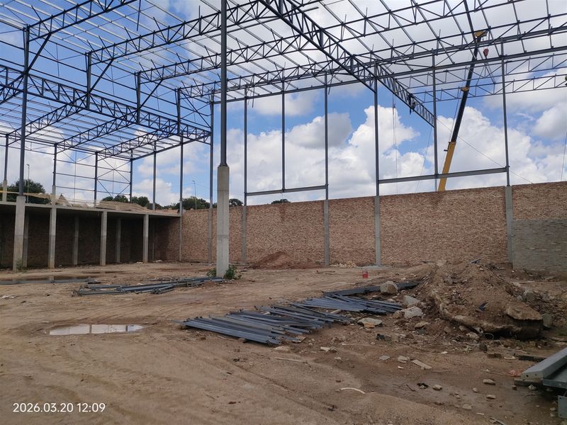 An industrial construction site featuring an exposed steel truss roof structure, steel support columns, and unfinished brick perimeter walls on a dirt foundation.