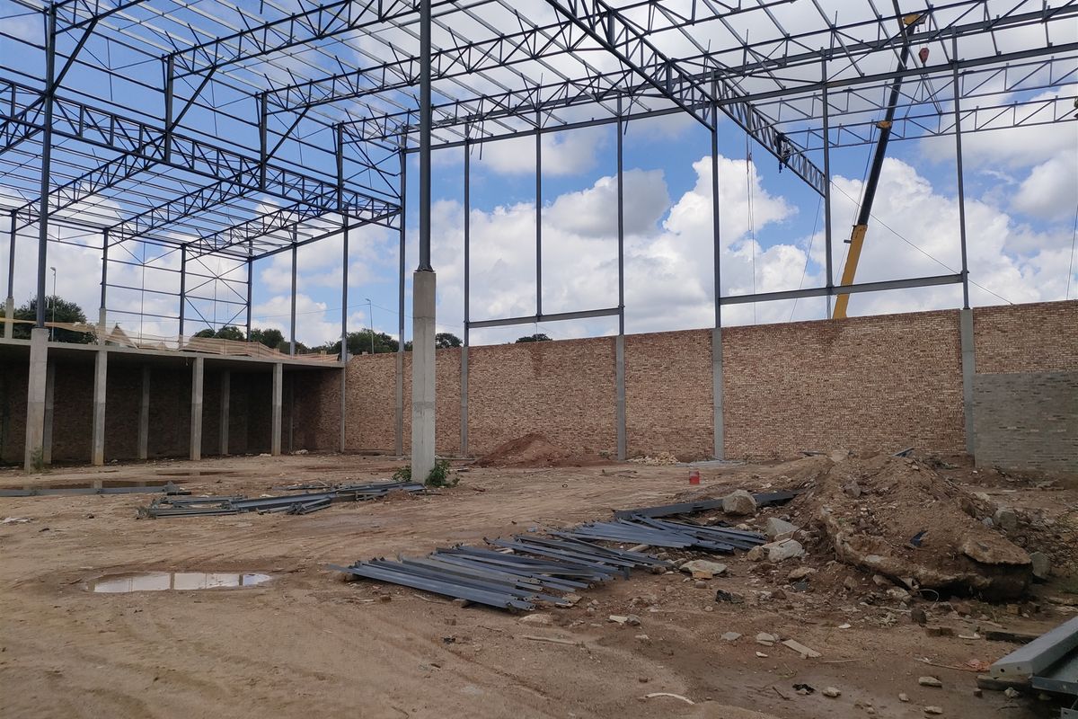 An industrial construction site featuring an exposed steel truss roof structure, steel support columns, and unfinished brick perimeter walls on a dirt foundation.
