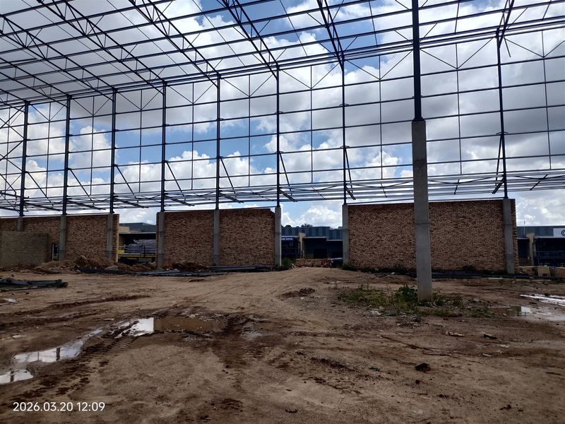 An industrial warehouse space under construction featuring a steel portal frame structure, exposed brick walls, and an unpaved earth floor.
