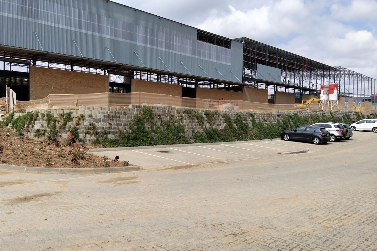 Exterior view of an industrial property under construction, featuring a large steel frame structure, metal cladding, brickwork, and a paved parking area.