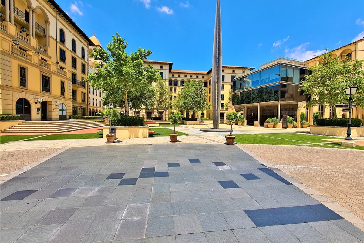 A well-maintained outdoor courtyard area within an office complex, featuring paved walkways, landscaped garden beds, and a central architectural sculpture.