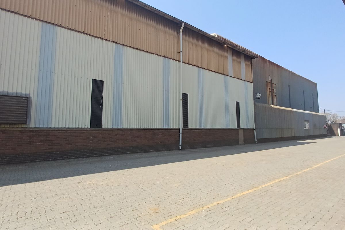 Exterior view of an industrial warehouse building featuring corrugated metal cladding, a brick base, and a paved driveway area.
