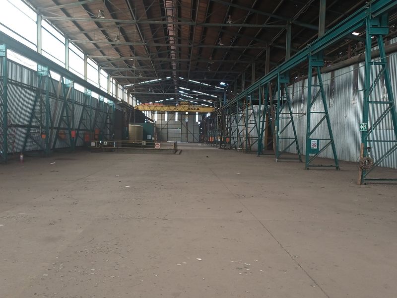 Expansive industrial warehouse space featuring high steel truss ceilings, corrugated metal walls, concrete flooring, and an integrated overhead gantry crane system.