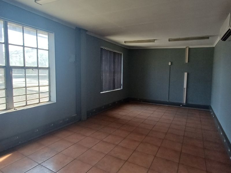 An empty, functional office space featuring tiled flooring, large steel-framed windows with vertical blinds, and a wall-mounted air conditioning unit.