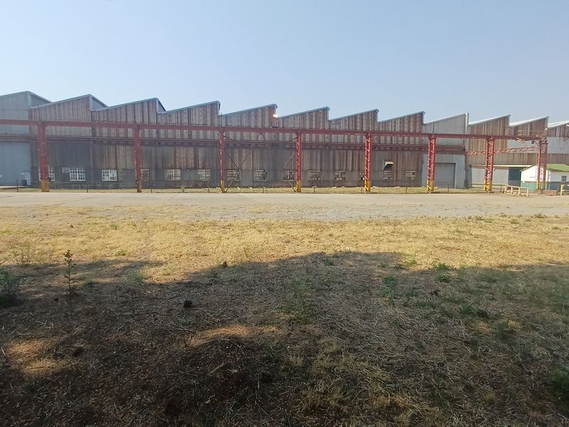 Large industrial warehouse facility featuring a sawtooth roof design, steel structural framework, and expansive concrete yard space.