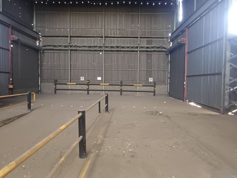Large industrial utility space featuring corrugated metal walls, high ceilings, roller shutter doors, and durable concrete flooring, suitable for storage or workshop operations.