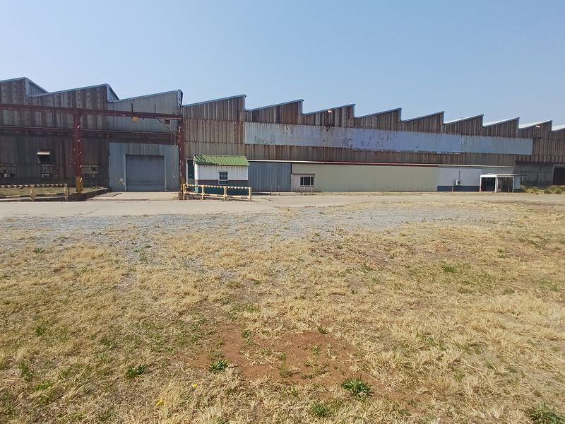 Large industrial warehouse facility featuring a sawtooth roof design, corrugated metal cladding, and a spacious concrete yard area.