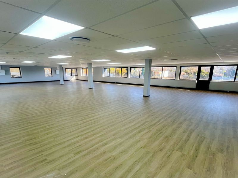 A spacious, open-plan office area featuring modern laminate flooring, a suspended ceiling with recessed lighting, and large windows providing ample natural light.