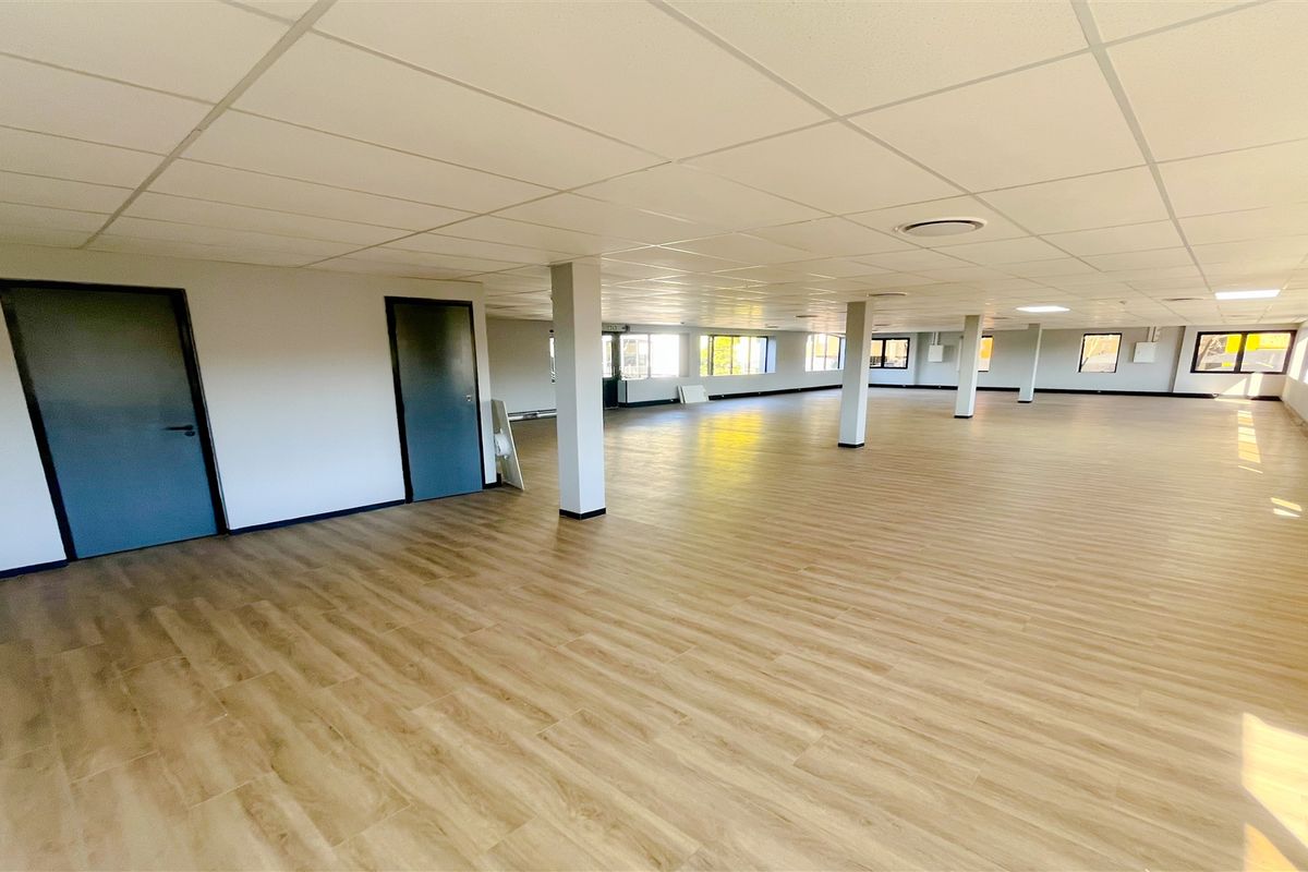 A spacious, open-plan office area featuring modern wood-look flooring, a suspended ceiling, and ample natural light from large windows.