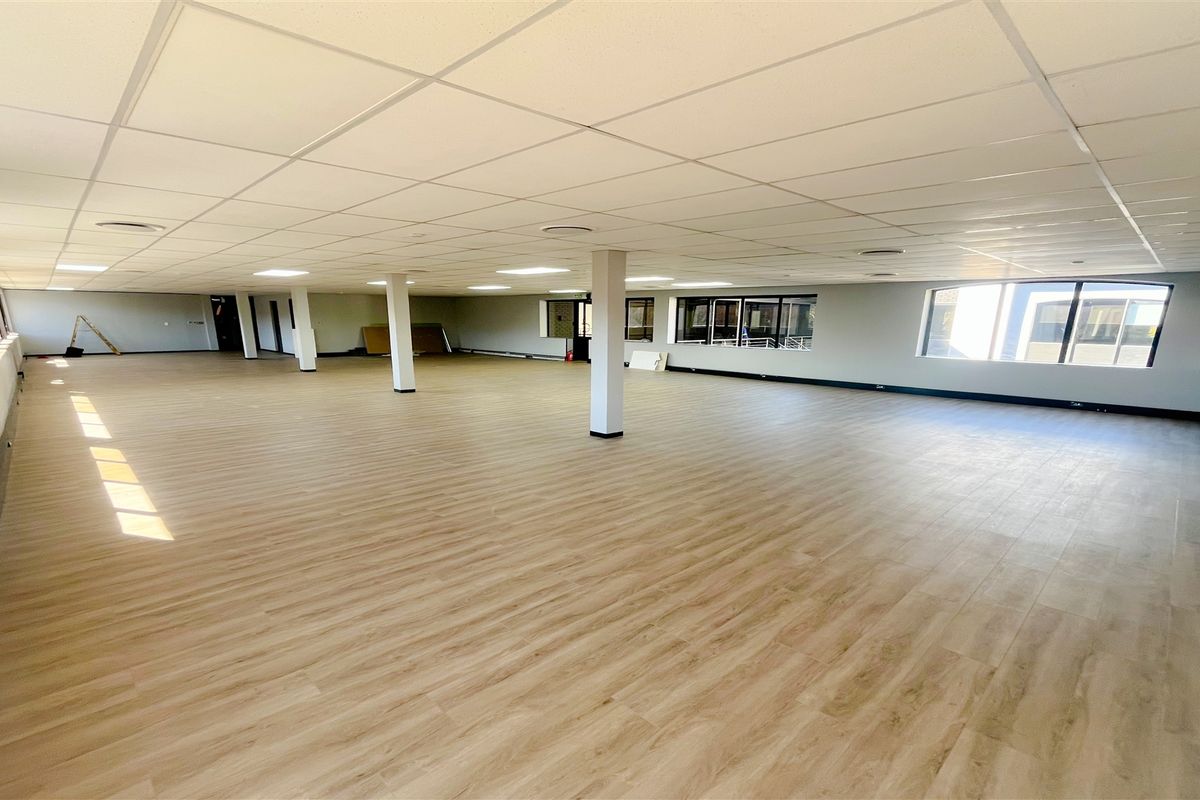A spacious, open-plan office area featuring modern laminate flooring, a suspended ceiling with recessed lighting, and large windows providing ample natural light.