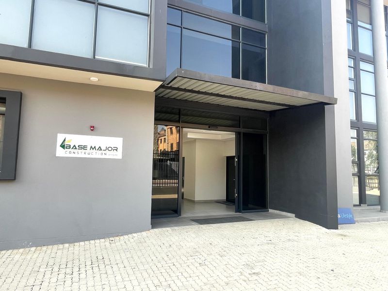 Modern office building entrance featuring a paved driveway, glass facade, and a metal canopy over the main sliding door access point.