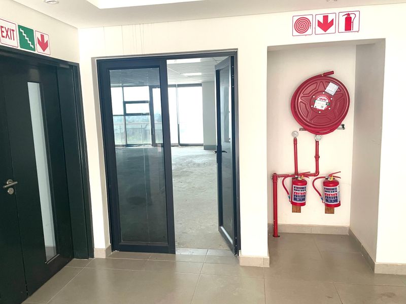 A clean office hallway featuring fire safety equipment, including a hose reel and extinguishers, with tiled flooring and access to an open office space.