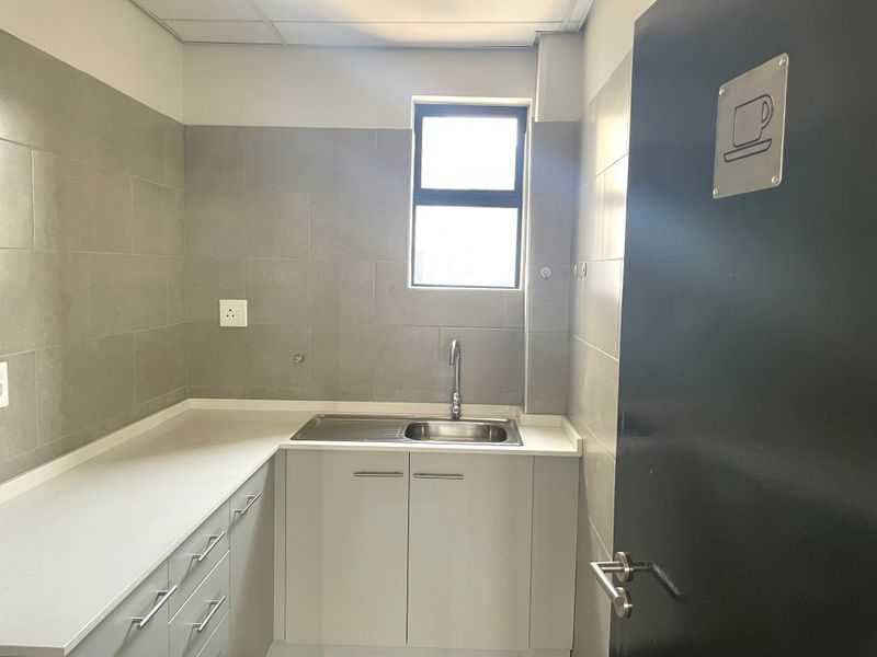 A clean and functional office kitchenette featuring modern grey cabinetry, a stainless steel sink, and neutral tiled walls.