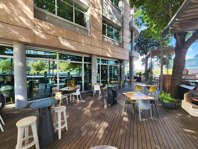 A spacious outdoor timber deck patio area featuring a pergola, integrated with the office building's glass facade and concrete structural columns.