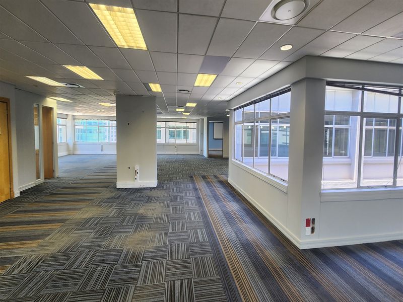 Spacious open-plan office area featuring suspended ceilings, recessed lighting, carpet tiles, and large windows providing ample natural light.