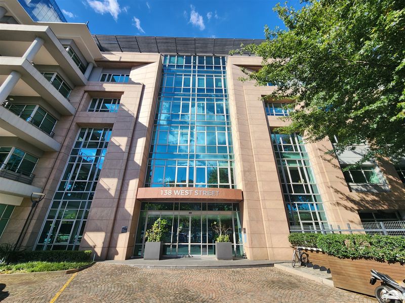 Modern commercial office building facade featuring a prominent glass curtain wall entrance, stone cladding, and a paved cobblestone driveway.