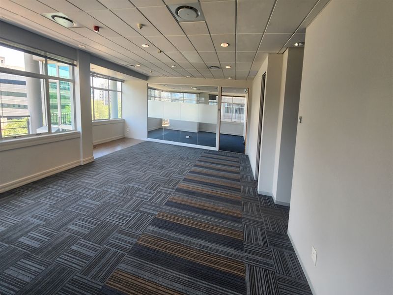 A bright, open-plan office space featuring carpet tiles, a suspended ceiling with recessed lighting, and a glass partition wall.