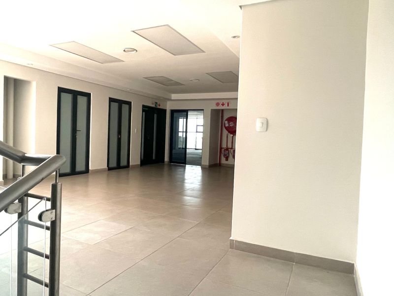 A spacious, well-lit office hallway featuring neutral tiled flooring, recessed ceiling lighting, and multiple access doors, including integrated fire safety equipment.