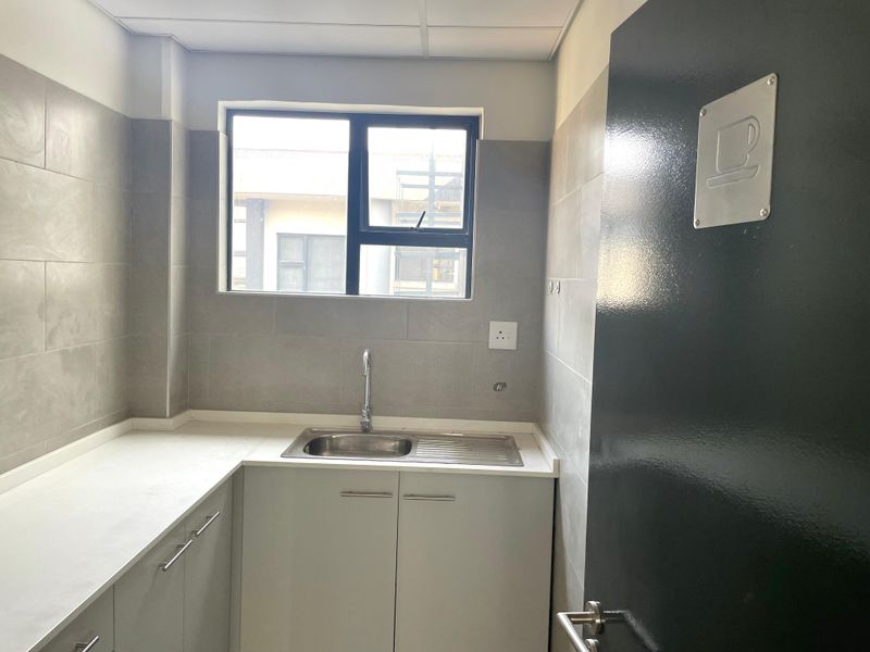 A clean, modern office kitchenette featuring grey tiled walls, built-in cabinetry, a stainless steel sink, and a neutral countertop.