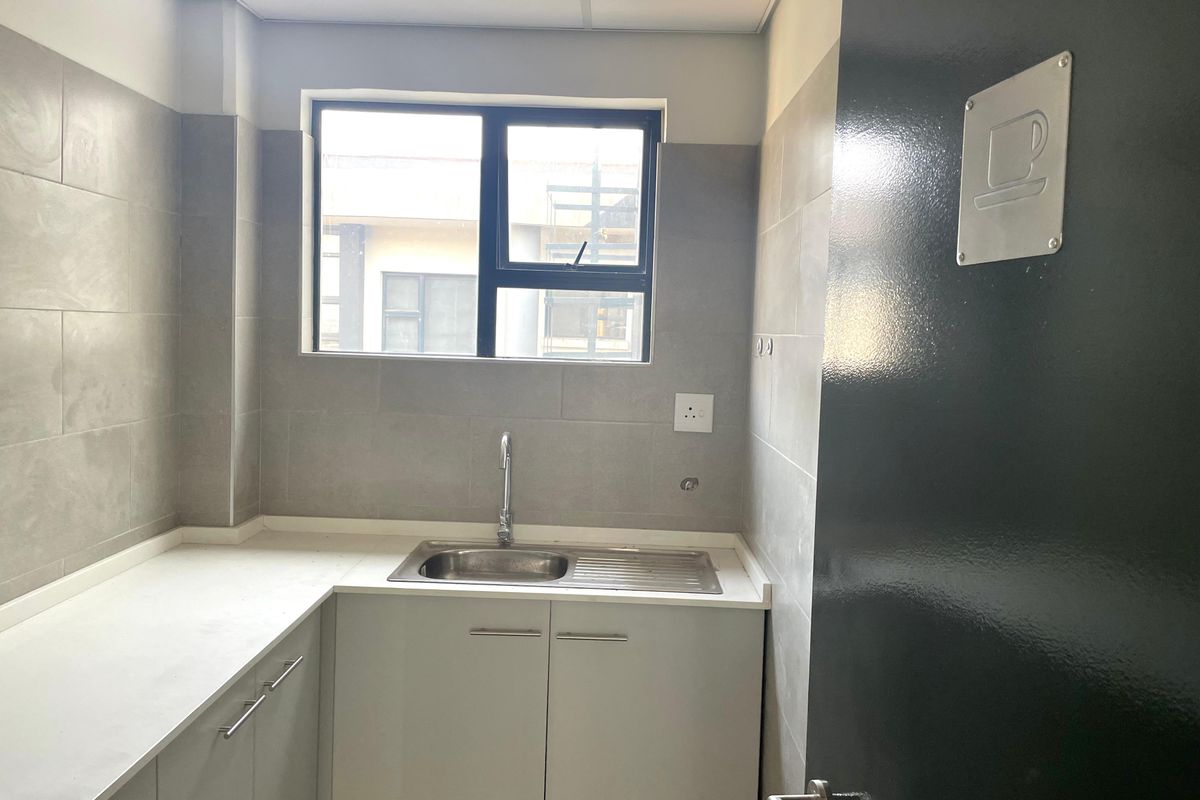 A clean, modern office kitchenette featuring grey tiled walls, built-in cabinetry, a stainless steel sink, and a neutral countertop.