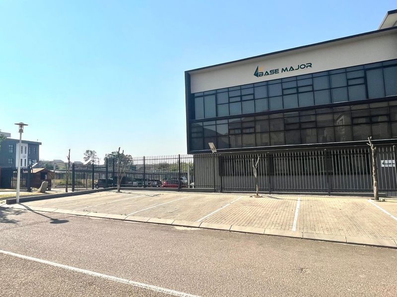 Front exterior view of a modern multi-story office building featuring a paved parking area and secure metal perimeter fencing.