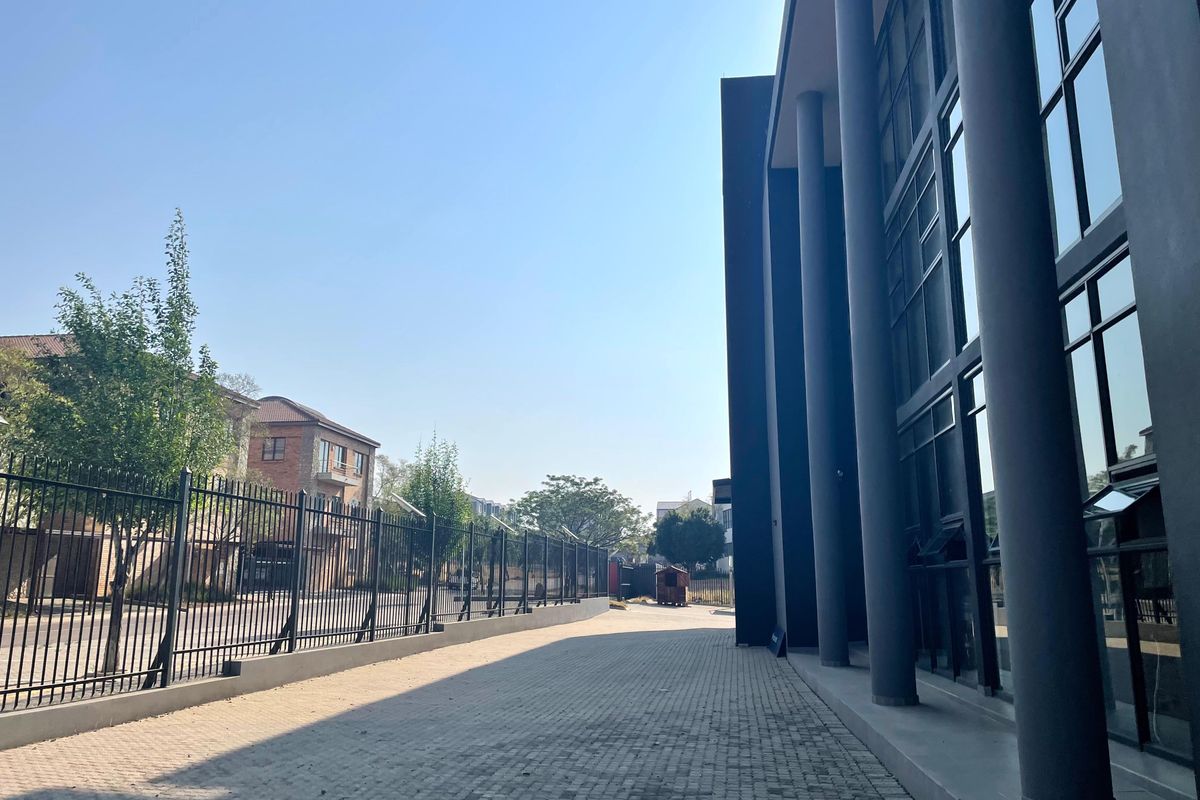 A modern office building exterior featuring a paved driveway, structural columns, and a large glass facade enclosed by metal perimeter fencing.