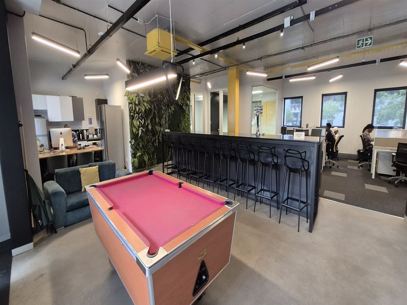Modern open-plan office space featuring a built-in bar counter, vertical garden wall, exposed industrial ceiling, and a mix of polished concrete and carpeted flooring.