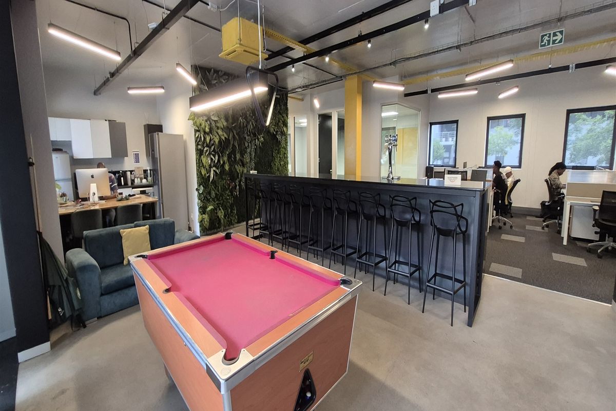 Modern open-plan office space featuring a built-in bar counter, vertical garden wall, exposed industrial ceiling, and a mix of polished concrete and carpeted flooring.
